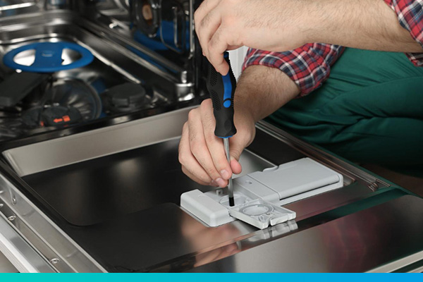 Zwei Hände in der Nahaufnahme, die an einer geöffneten Geschirrspülertür mit einem Schraubendreher schrauben