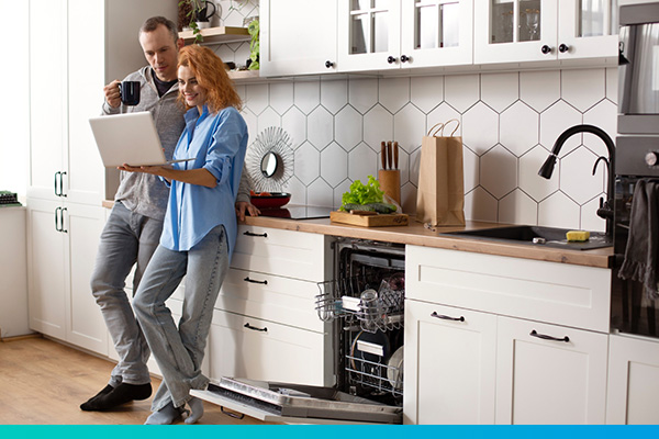 Paar mit Laptop in der Hand steht in einer Küche neben geöffneten Geschirrspüler