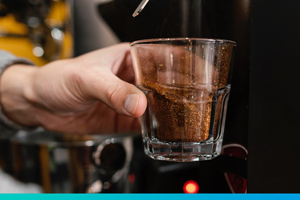 Eine Hand hält ein Glas mit Kaffeepulver vor einer Kaffeemaschine