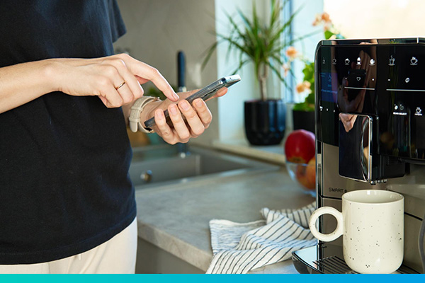 Nahaufnahme: Oberkörper einer Frau, die auf einem Handy tippt, vor einer Kaffeemaschine