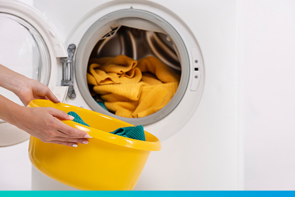 Zwei Hände halten eine Schüssel mit Wäsche vor einen geöffneten Wäschetrockner, in dem sich auch Wäsche befindet.