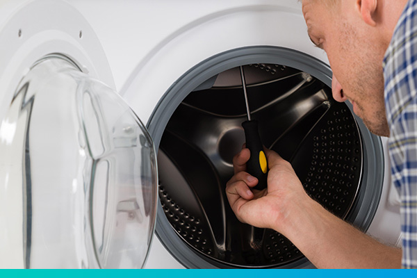 Nahaufnahme einer geöffneten Wäschertrocknertür mit einem Techniker davor, der einen Schraubendreher in die Maschine hält.